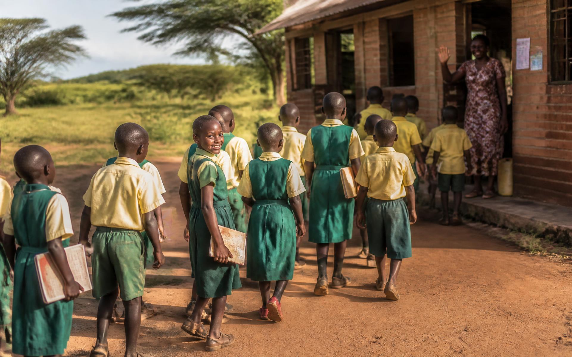 Schoolchildren in uniform walking together with their books as a teacher welcomes them outside a rural classroom